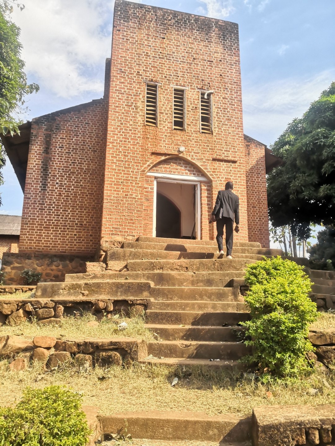 A man in a suit carrying copies of The Prayer walks up the stone steps into a red-brick church in rural Malawi