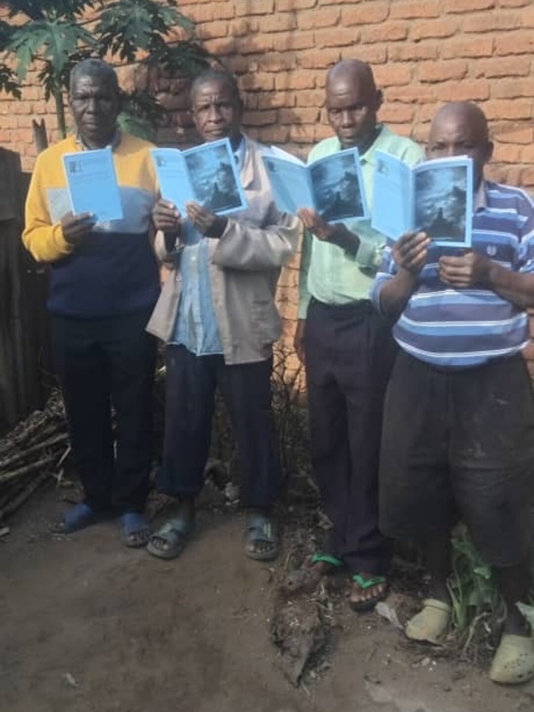 Four men in rural Malawi standing against a brick wall, each reading a copy of Pemphero, the Chichewa edition of The Prayer