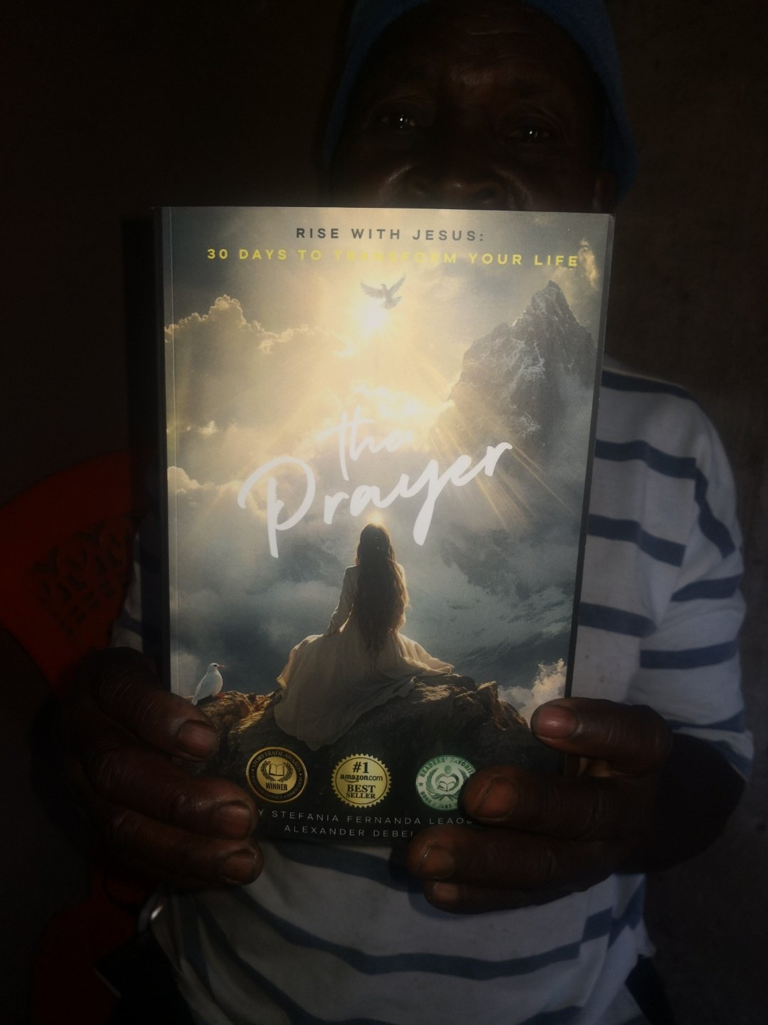 An elderly man in Malawi holding the paperback of The Prayer up to the camera, the glowing cover and award seals visible