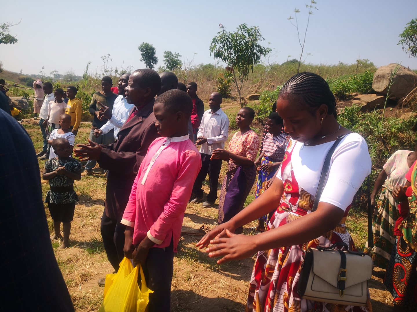 Men, women and children in Malawi worshipping together outdoors in the open air, hands clapping, beneath a clear sky