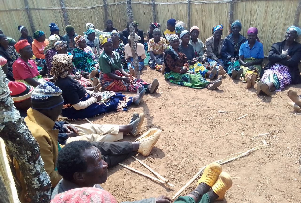 Dozens of women in bright headscarves and chitenje wraps seated together on the ground inside a reed-walled meeting place in Malawi