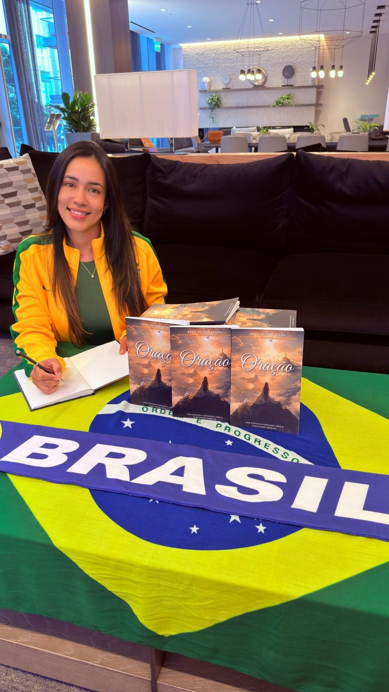 Stefania Leão in a yellow Brazil jacket signs a copy of Oração beside three copies fanned across the Brazilian flag
