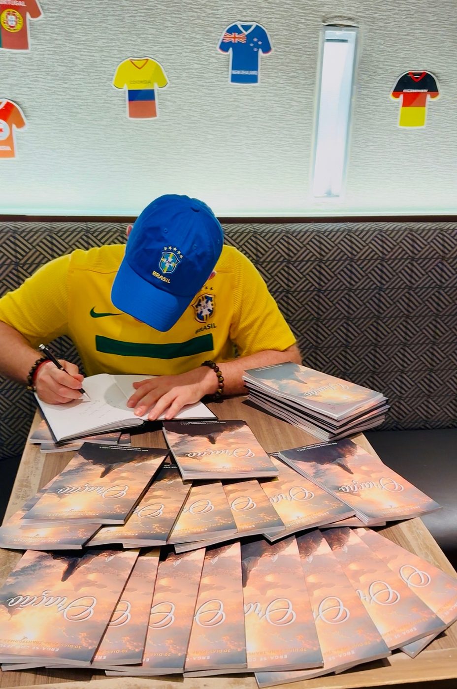 Alexander Debelov signing a large stack of Oração copies beneath a wall of World Cup national-team jerseys