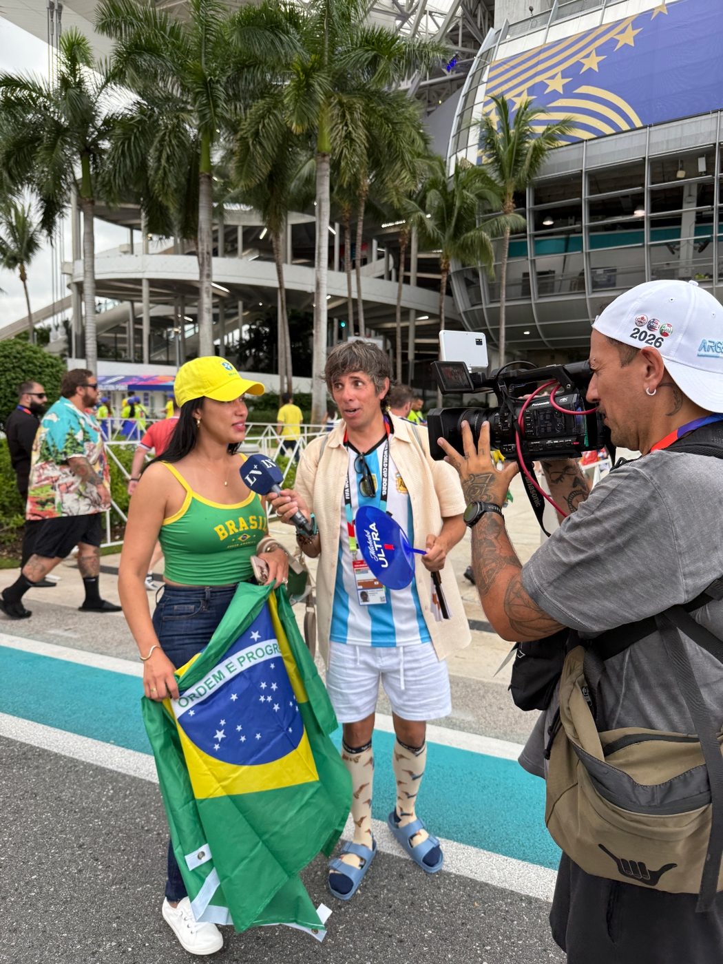 Stefania Leão in a Brazil top and yellow cap, interviewed by a TV crew outside the stadium with a Brazilian flag