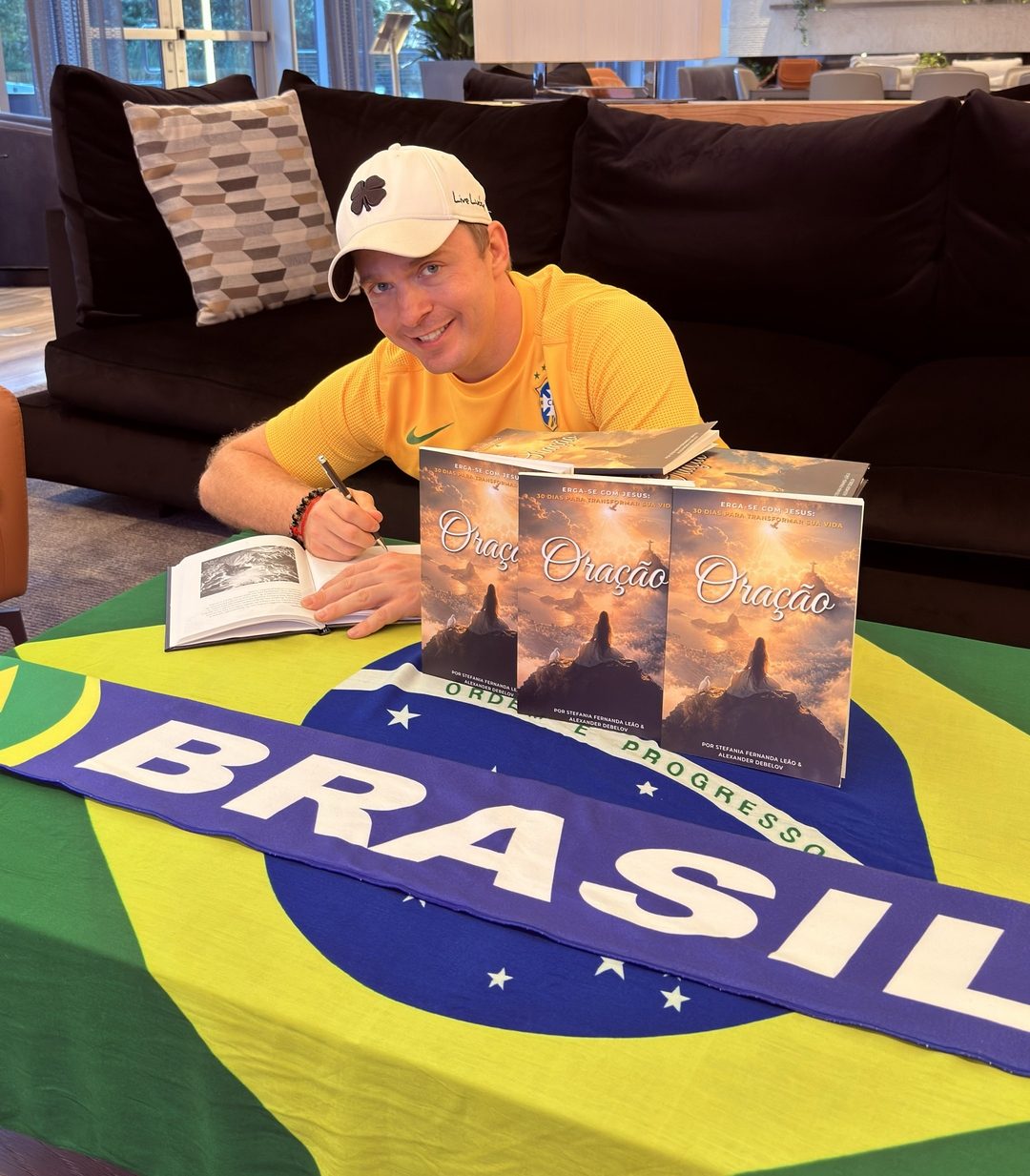 Alexander Debelov in a yellow Brazil jersey signing copies of Oração on a Brazil flag tablecloth.