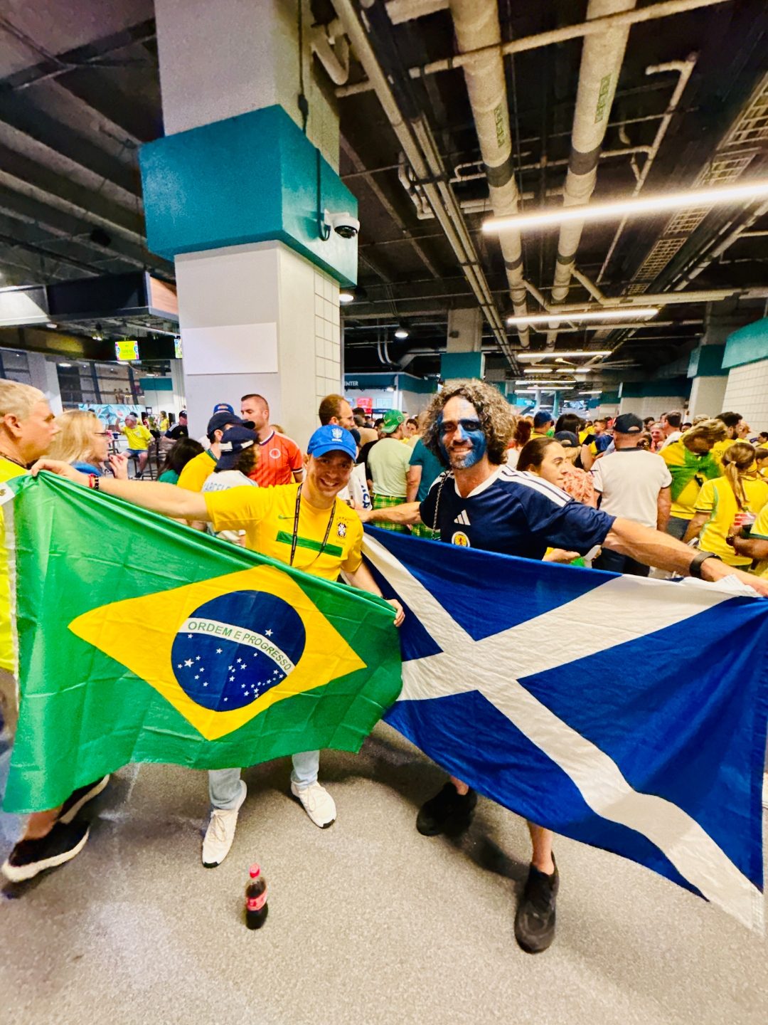 Alexander Debelov holding the Brazilian flag beside a Scotland supporter with painted face and Scotland flag inside the World Cup stadium concourse.